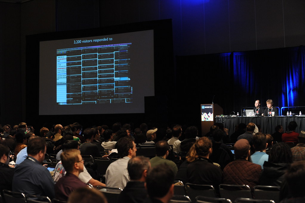 Attendees fill a conference hall at the Game Developers Conference, with rows of seats and large presentation screens visible.